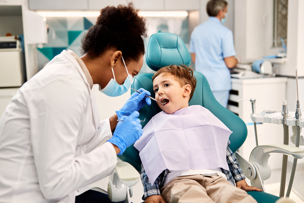 Black female dentist examining small boy's teeth during dental procedure at dentist's office. Focus is on boy.
