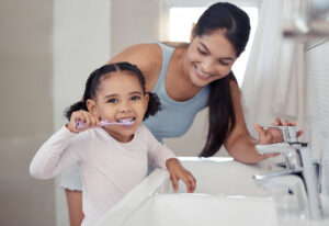 Brushing teeth, girl or mother teaching learning child personal hygiene, dental care or healthcare in house or family home bathroom. Happy smile woman, parent and kid in wellness with sink toothbrush.